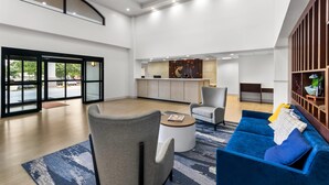Eine geräumige Lobby mit einem blauen Sofa, grauen Sesseln, einem runden Couchtisch und einer Küchenzeile im Hintergrund.