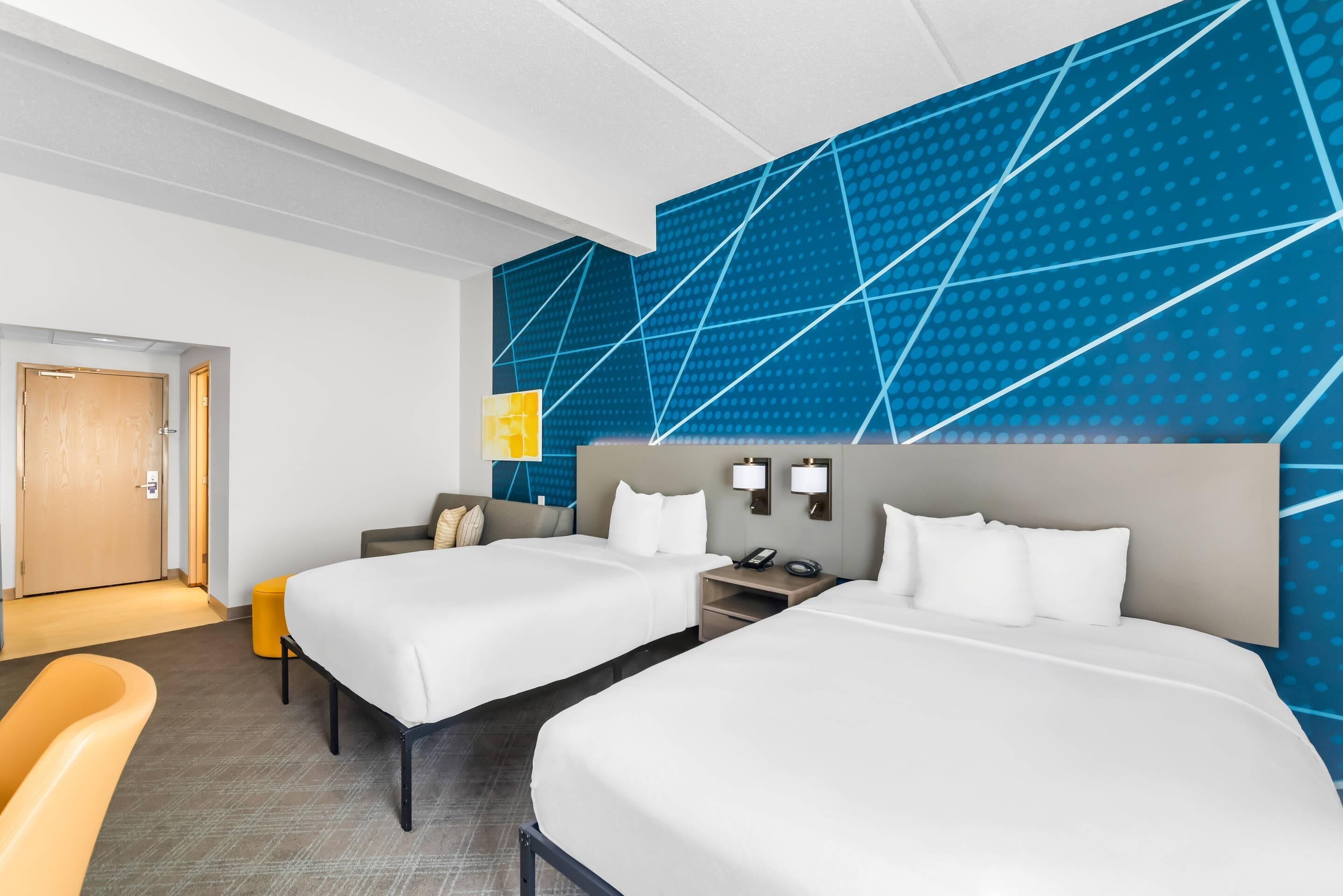 A hotel room with two beds, a desk, a chair, a nightstand, and a wall with a geometric pattern.
