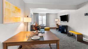 Ein Hotelzimmer mit Esstisch für zwei Personen, einer Couch, einem Fernseher und einem Fenster mit Vorhängen.