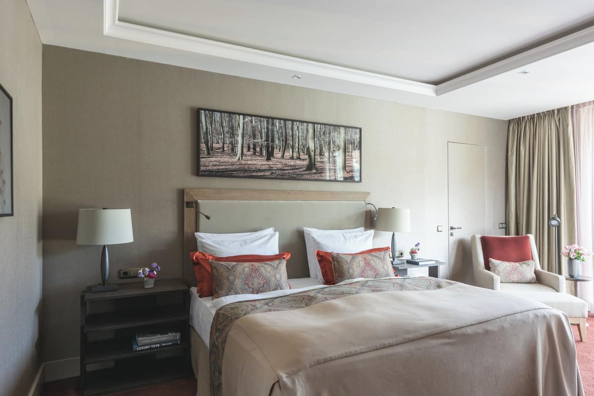 Ein Hotelzimmer mit einem großen Bett, zwei Nachttischen, einem Sessel, einem Bild an der Wand und einem Beistelltisch mit einer Lampe.