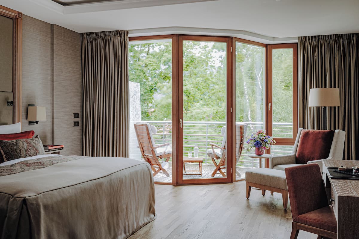 Ein Hotelzimmer mit einem großen Bett, einem Schreibtisch mit Lampe, einem Sessel, einem kleinen Tisch mit Blumenvase und Blick auf Grün hinter einer Glastür.