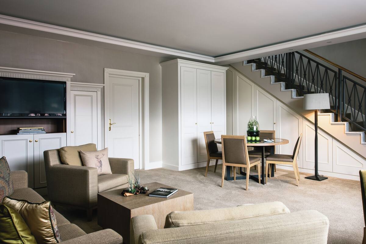 Ein Wohnzimmer mit einer Couch, Couchtisch und einem Essbereich mit Stühlen und Tisch. Ein Fernseher ist an der Wand montiert, eine Treppe mit Geländer und eine Tür mit Griff sind ebenfalls vorhanden.