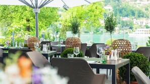 An outdoor restaurant with tables set for dining, surrounded by greenery and a view of a body of water. - Althoff Seehotel Überfahrt (Rottach-Egern)
