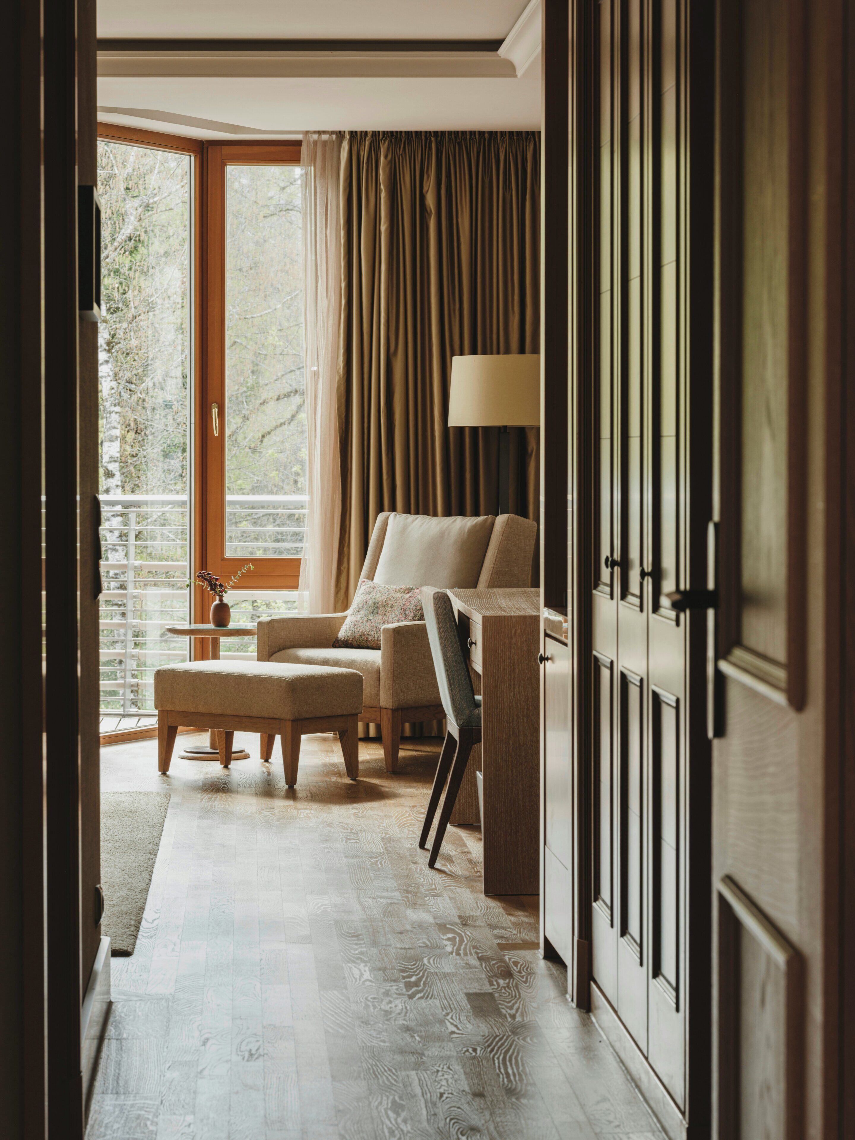 A room with a sofa, armchair, and ottoman, a lamp, and a view through a window with curtains.