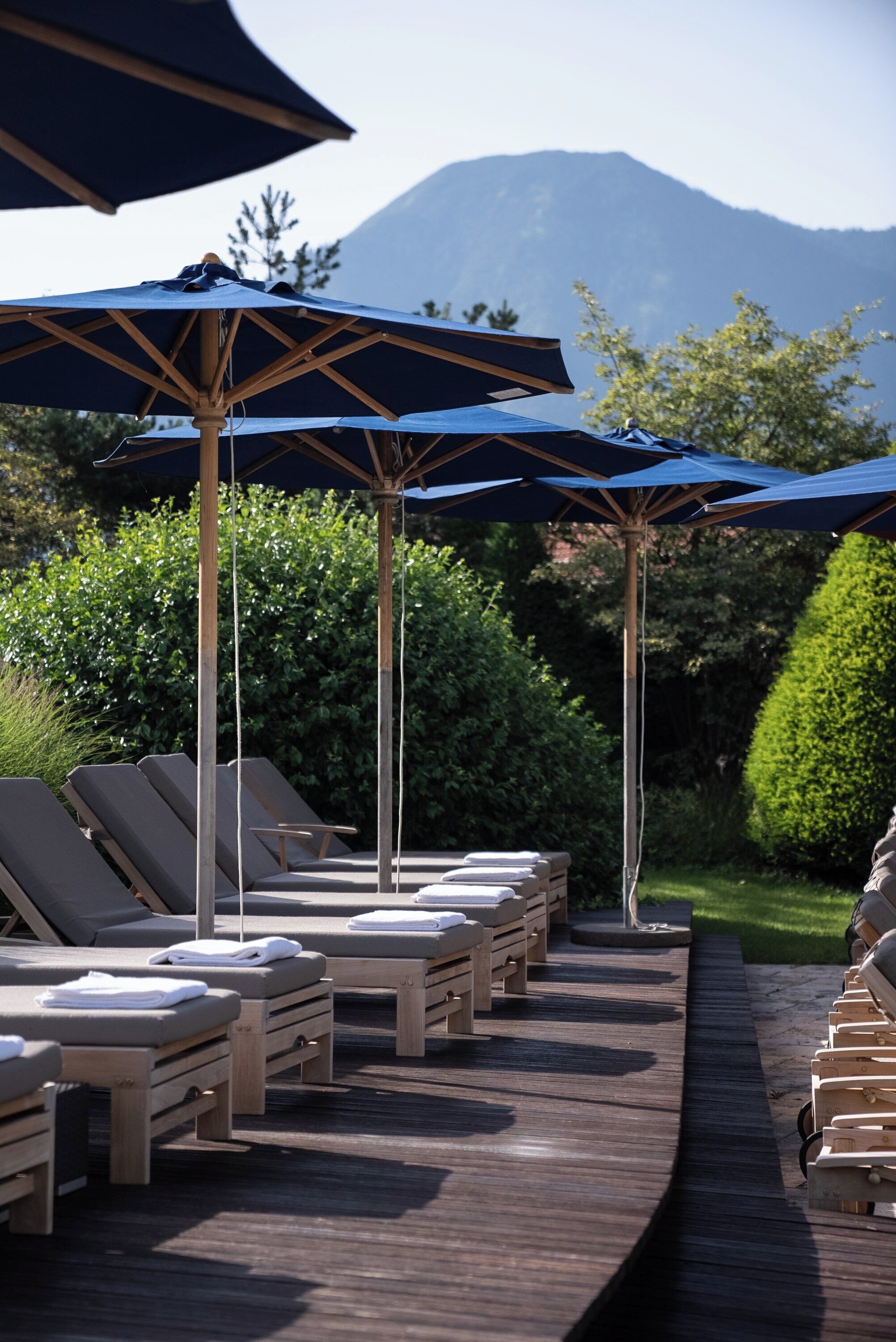 Ein Poolbereich mit Liegestühlen und Sonnenschirmen, ein Holzdeck und ein Berg im Hintergrund.