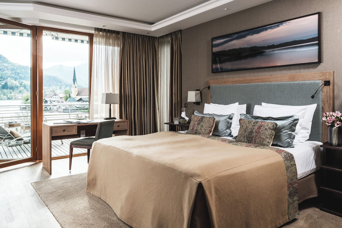 Ein Hotelzimmer mit einem großen Bett, einem Schreibtisch und einem Stuhl. Es gibt ein Fenster mit Vorhängen, ein Bild an der Wand sowie einen Blick auf Stadt und Berge.