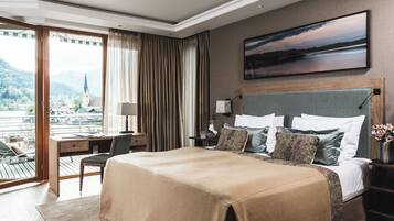 Quarto de hotel com uma cama grande, uma escrivaninha e uma cadeira. Há uma janela com cortinas, um quadro na parede e vista para uma cidade e montanhas.