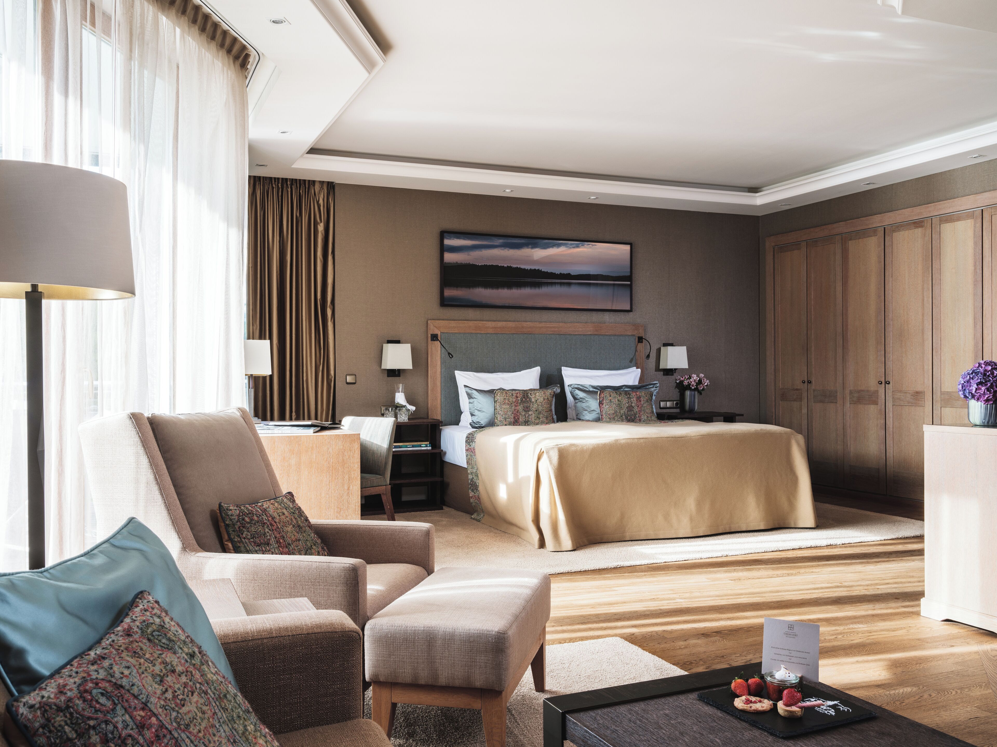 A hotel room with a large bed, a sitting area with a sofa and armchairs, a desk, and a view of the outdoors through the window.
