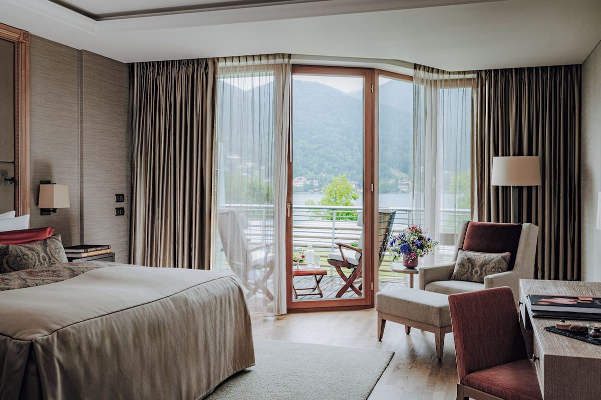 Ein Hotelzimmer mit Bett, Schreibtisch, Stuhl und Blick auf einen See und Berge durch eine große Glasschiebetür.