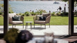 A room with a large window overlooking a lake and mountains, furnished with wicker chairs and a small table. - Althoff Seehotel Überfahrt (Rottach-Egern)