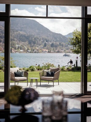 A room with a large window overlooking a lake and mountains, furnished with wicker chairs and a small table. - Althoff Seehotel Überfahrt (Rottach-Egern)