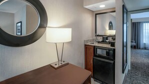 A hotel room with a wooden desk, a round mirror, a wall-mounted lamp, a kitchenette with a microwave, oven, and sink. - Embassy Suites by Hilton Nashville at Vanderbilt (Nashville)