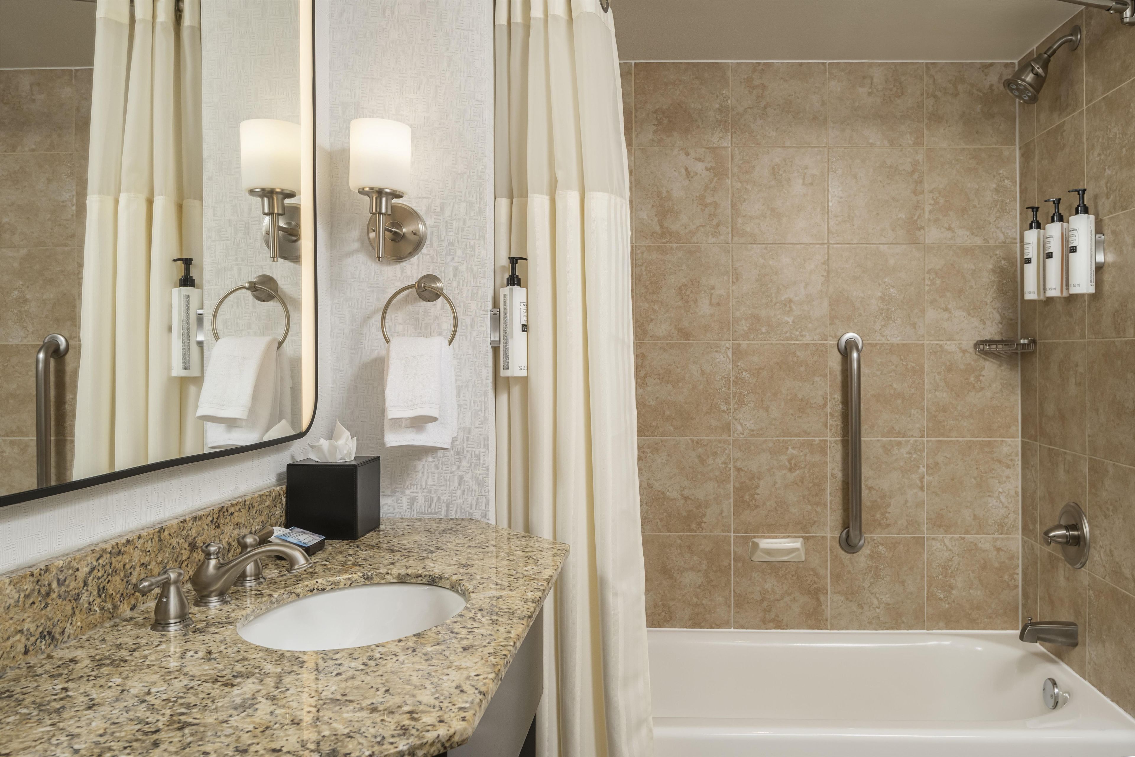A bathroom with a granite countertop sink, a mirror, a towel rack, a shower with a handheld showerhead, and a grab bar.