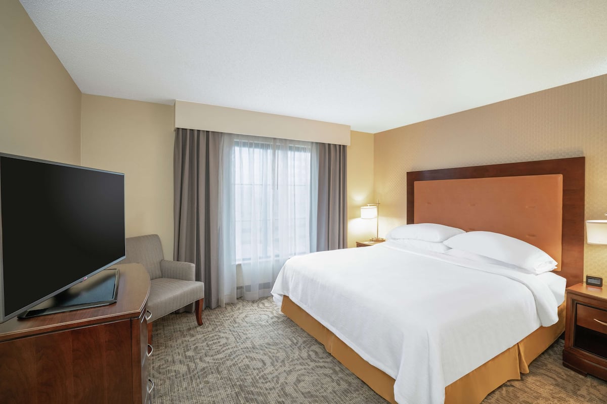 Una habitación de hotel con una cama grande, un televisor sobre un mueble de madera, una silla, un escritorio con lámpara y una ventana con cortinas.