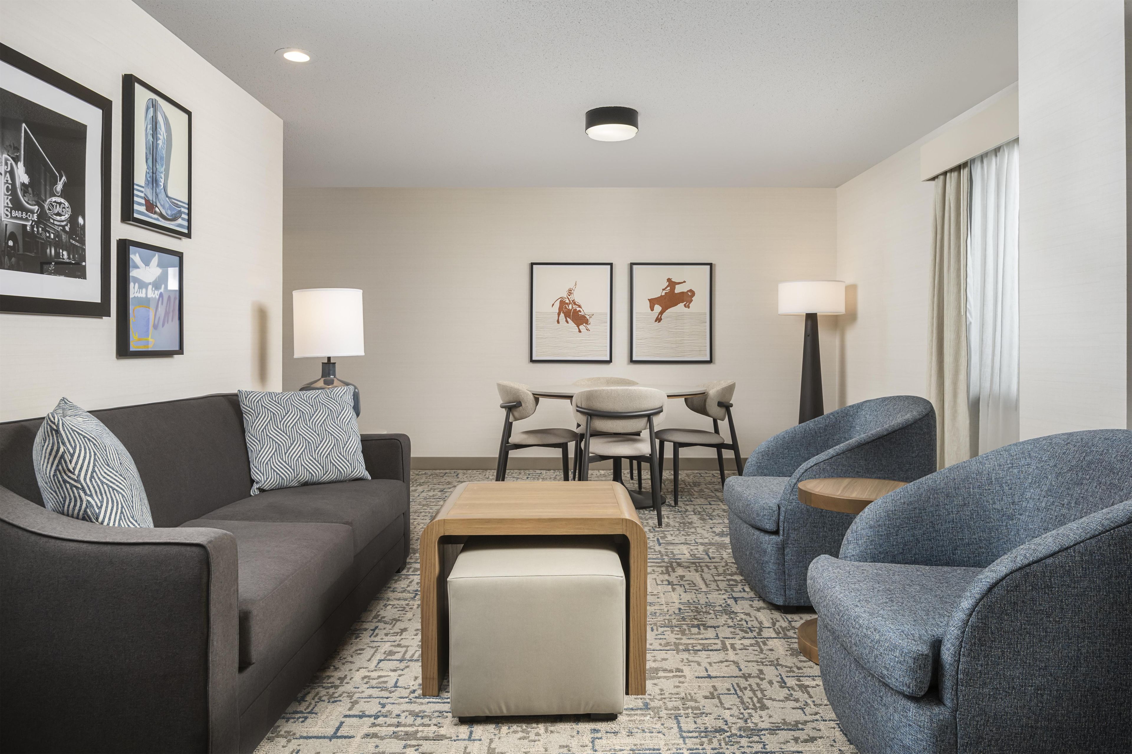 Una habitación de hotel moderna con un sofá gris, una pequeña mesa de madera, dos sillones, una mesa redonda con sillas y obras de arte enmarcadas en las paredes.