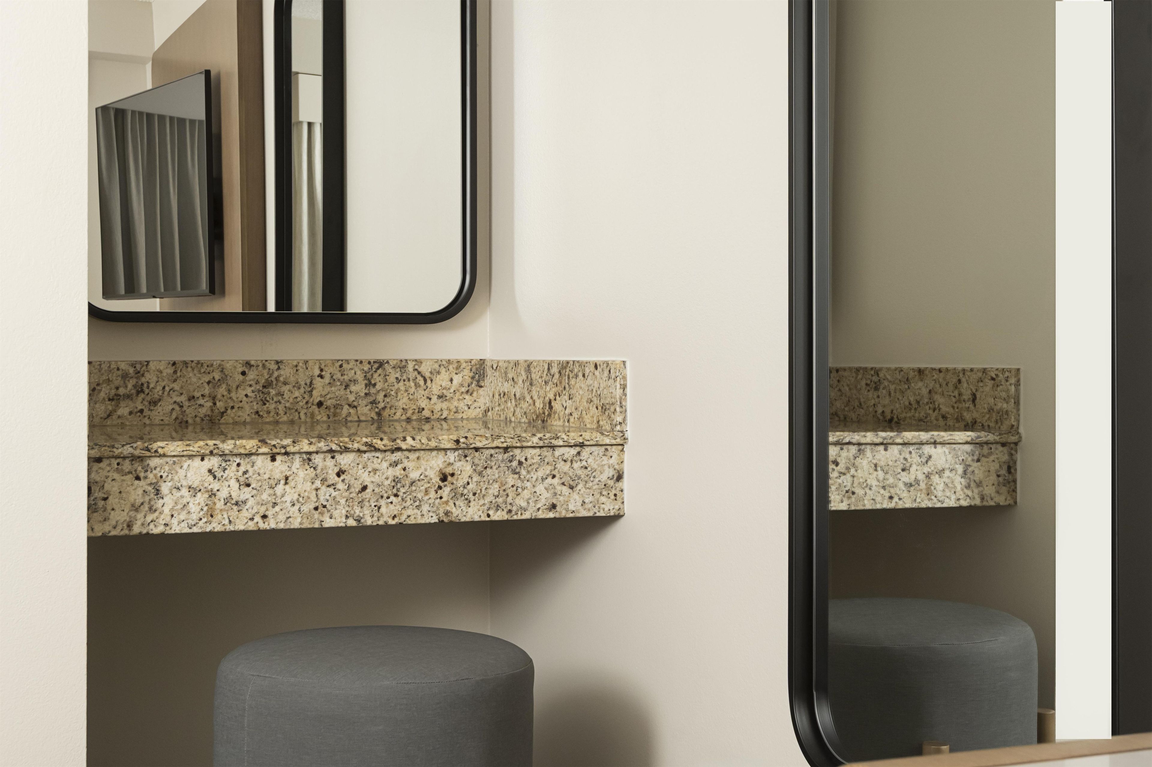 A bathroom with a granite countertop, a mirror, and a grey ottoman.