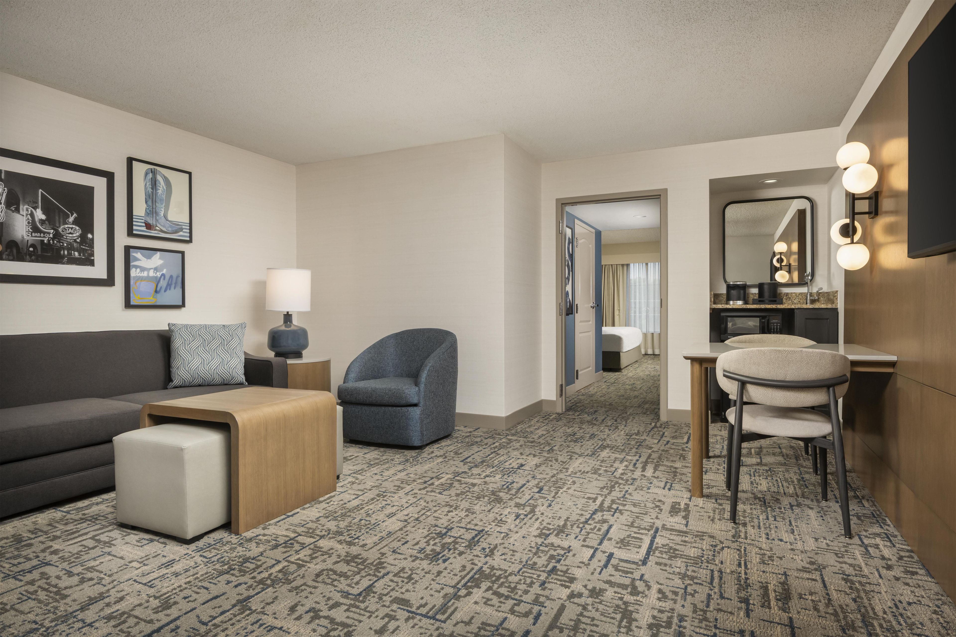 A hotel room with a sofa, armchair, ottoman, a small table, a lamp, a mirror with lights, and a carpeted floor.