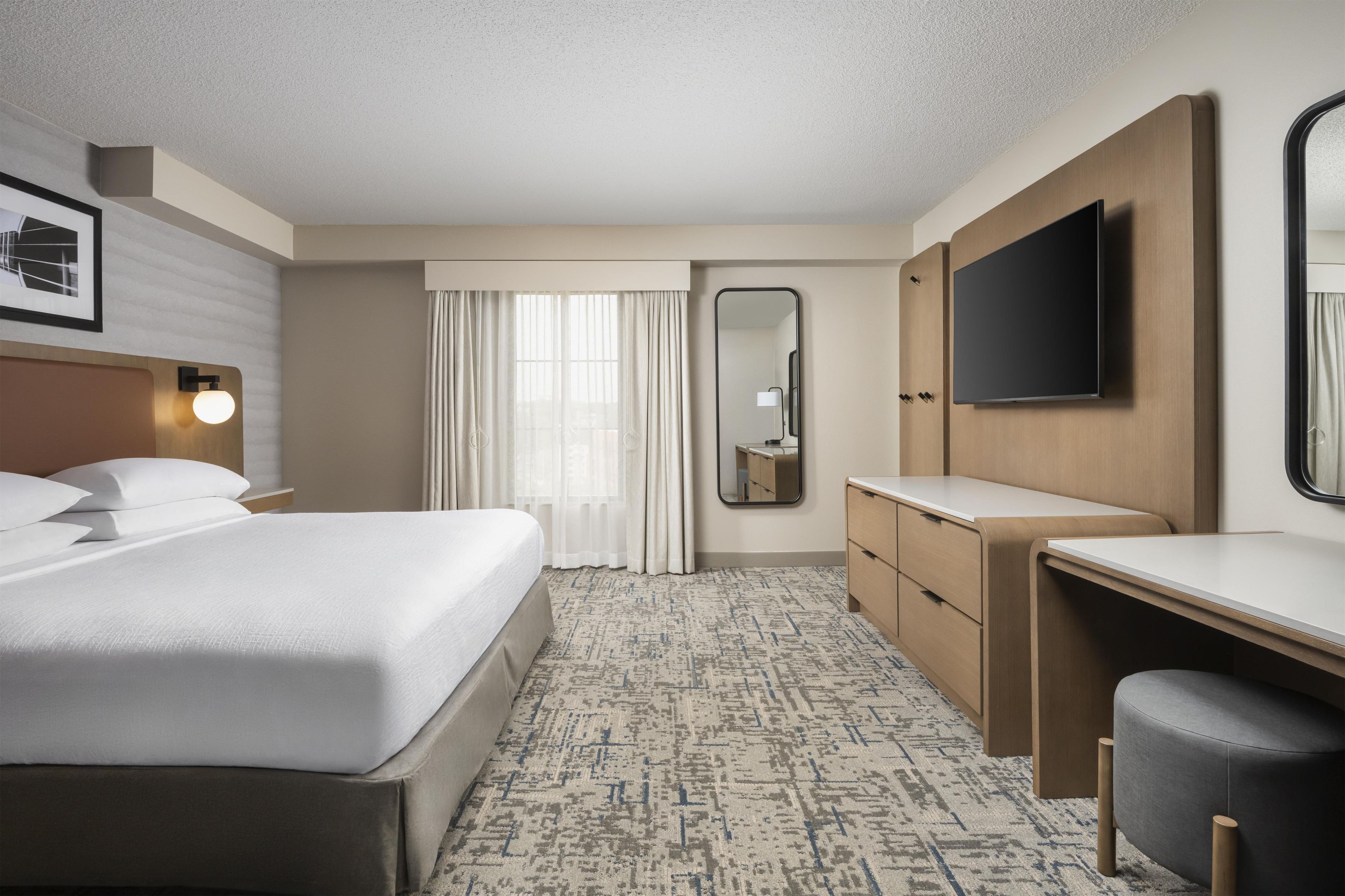 A hotel room with a bed, a desk, a chair, a TV, a mirror, and a window with curtains.