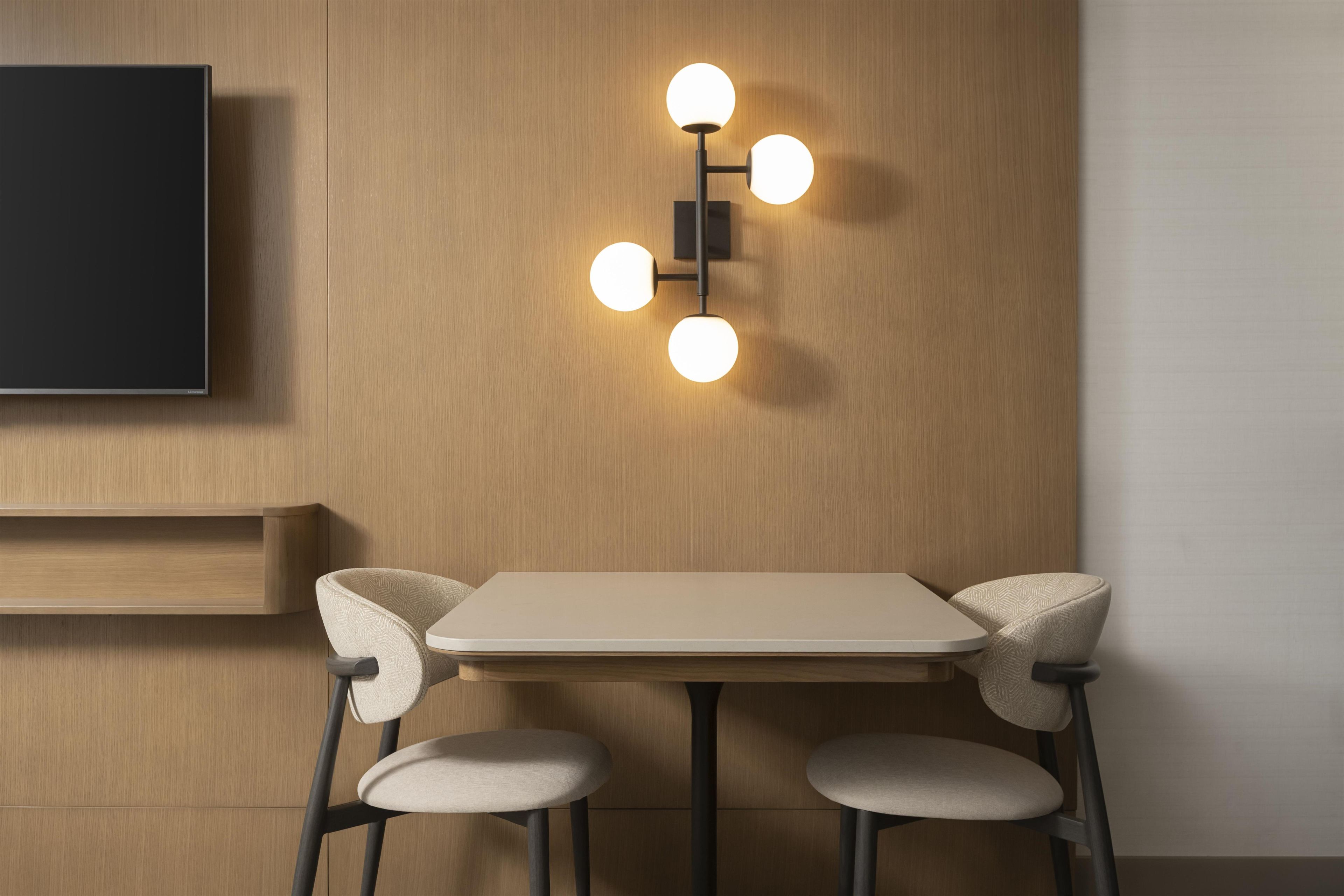 A modern room with a flat-screen TV mounted on the wall, a table with two chairs, and a wall-mounted light fixture with four bulbs.