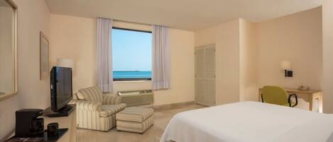 Habitación de hotel con cama, televisión, escritorio con silla, ventana con cortinas y vista al mar.
