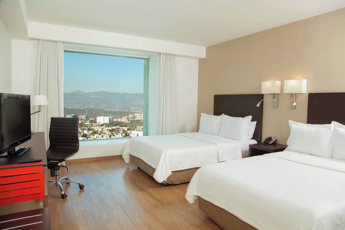 Habitación de hotel con dos camas, un escritorio, un televisor, una silla y vista a un paisaje urbano.