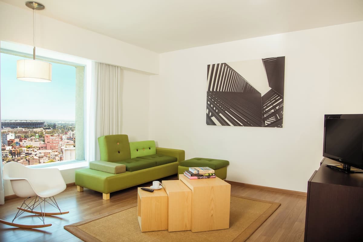 Una sala de estar moderna con un sofá verde, una mesa de centro de madera, una mecedora blanca y un amplio ventanal con vistas a la ciudad.