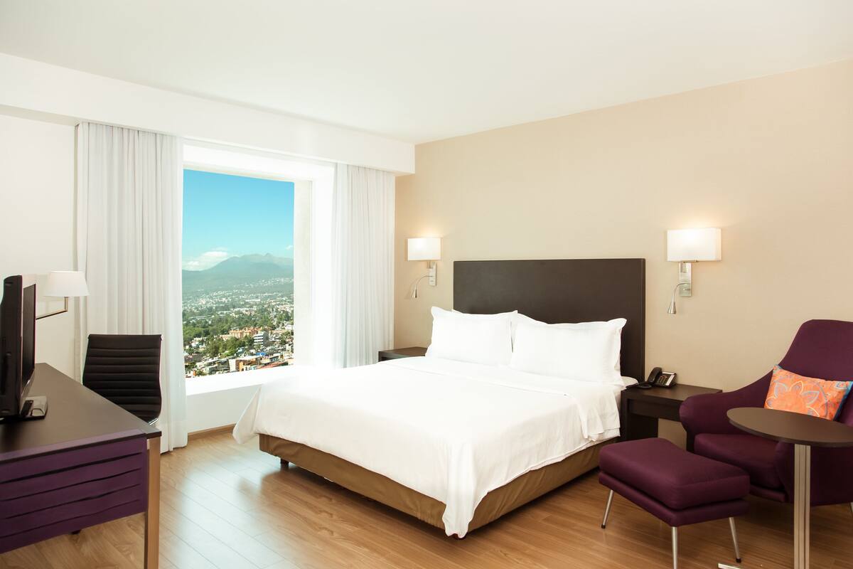 Habitación de hotel con cama, escritorio, silla, mesita y vista a la ciudad.