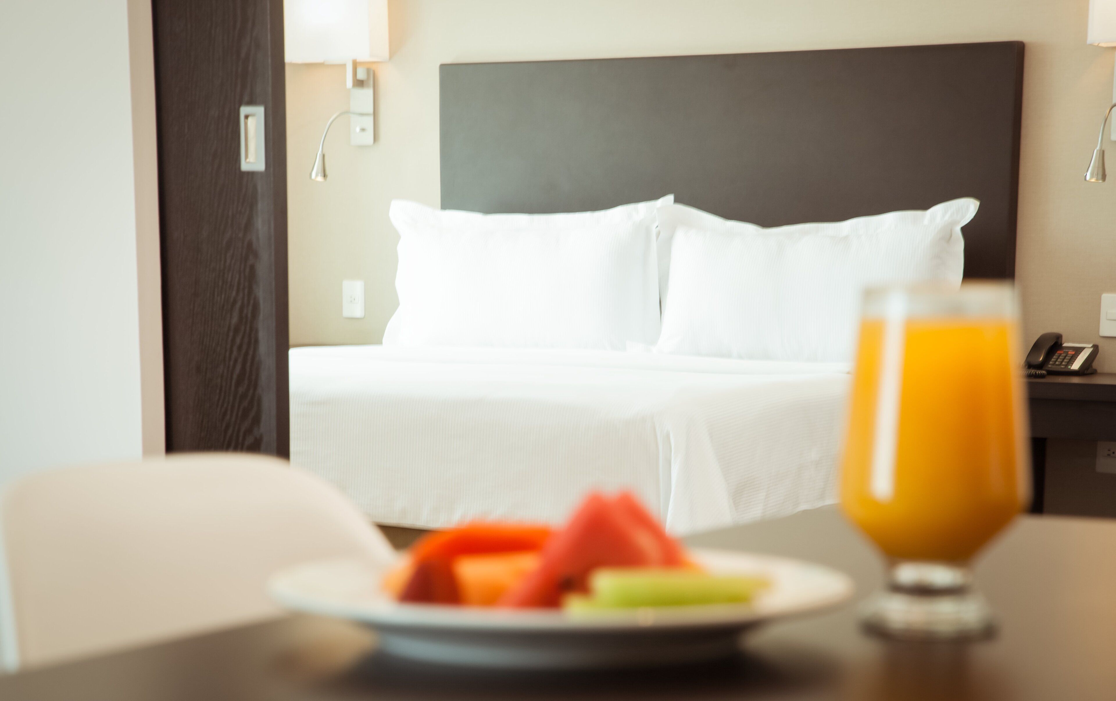A hotel room with a bed, a bedside table with a plate of fruit and a glass of orange juice, and a phone on the nightstand.