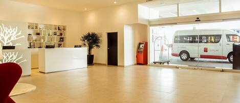 A hotel reception area with a reception desk, a plant, a self-service kiosk, and a van outside.