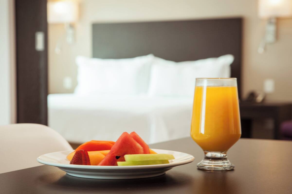 Habitación de hotel con una cama bien hecha, una mesita de noche con una bandeja de frutas y un vaso de zumo de naranja.
