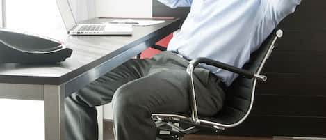 A man sitting in a modern office chair at a desk with a laptop, phone, and a red notebook.