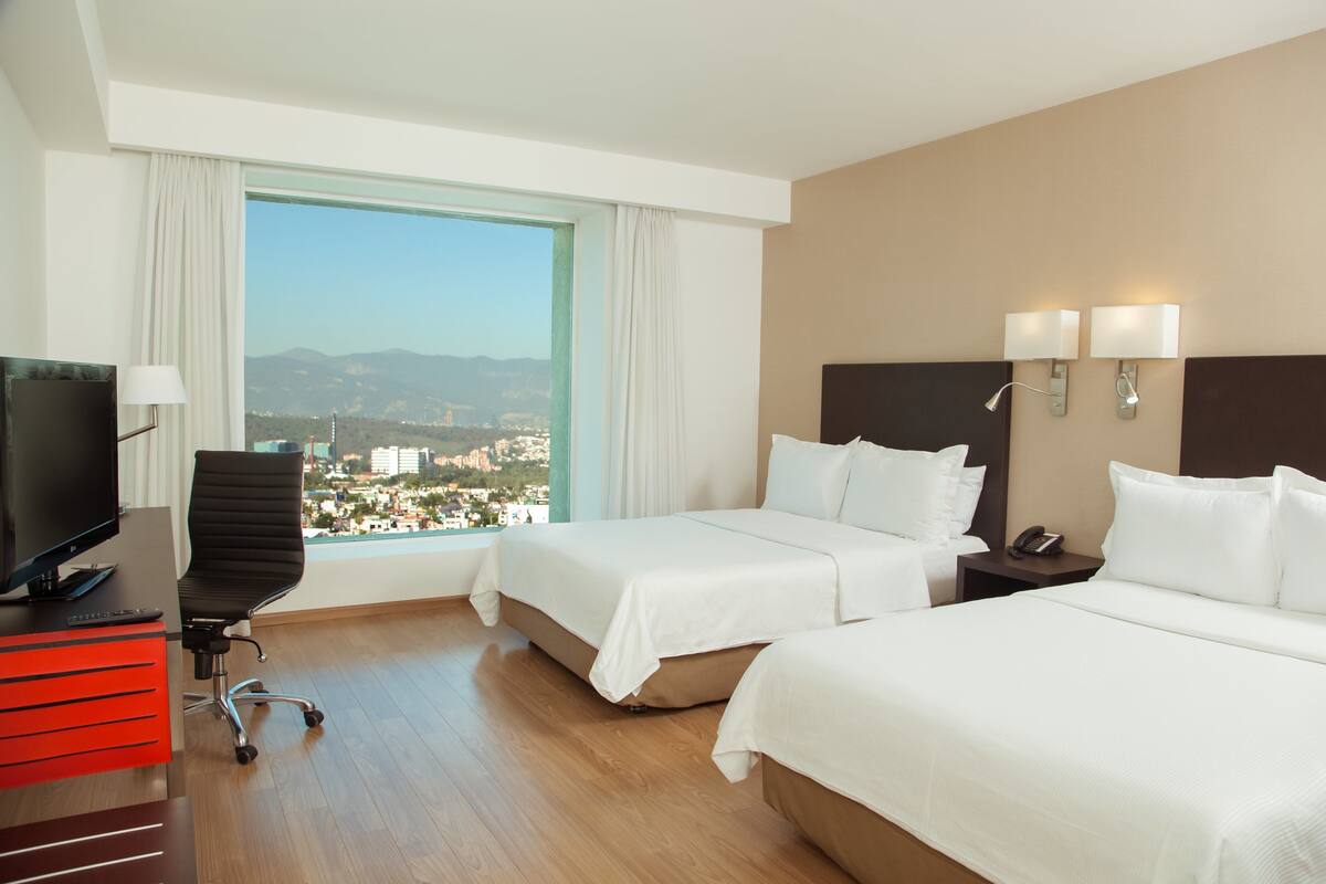 Habitación de hotel con dos camas, un escritorio con televisor, una silla y vistas a la ciudad.