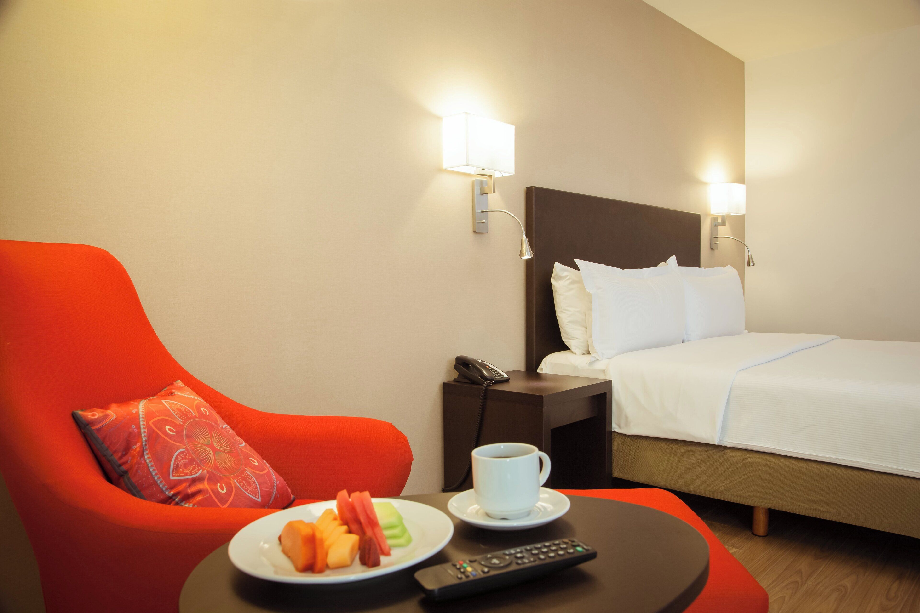A hotel room with a bed, a red armchair, a nightstand with a phone, a plate of fruit, a cup of coffee, and a remote control.