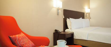 A hotel room with a bed, a red armchair, a nightstand with a phone, a plate of fruit, a cup of coffee, and a remote control.