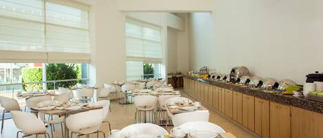 A spacious dining area with multiple tables and chairs, a long counter with various dishes, and large windows providing natural light.