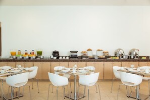 Un bufé de desayuno con diversos alimentos y bebidas, sillas blancas y una cocina moderna y limpia.