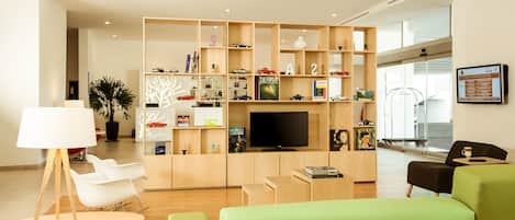 A modern hotel lobby with a wooden bookshelf, a flat-screen TV, and seating areas.