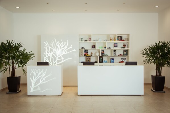 A reception area with a white reception desk, a bookshelf with various items, and two potted plants.