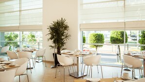 A modern restaurant with white chairs and tables, large windows, and a potted plant. - Fiesta Inn Periferico Sur (Mexico City)