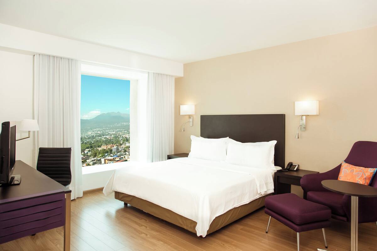 Habitación de hotel con una cama grande, un escritorio, una silla y vistas a un paisaje urbano a través de una ventana.