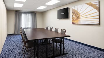 Une salle de conférence avec une longue table, des chaises, un téléviseur fixé au mur et une grande fenêtre avec des rideaux.