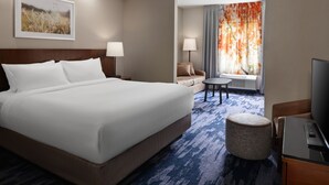 A hotel room with a large bed, two bedside tables, a TV, a small round ottoman, a sofa, a chair, and a window with curtains. - Fairfield Inn by Marriott Charlotte Mooresville (Mooresville)