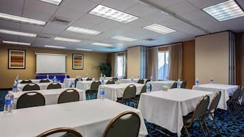 Un salón de conferencias con varias mesas, sillas y una pantalla para proyector. Las mesas están cubiertas con manteles blancos y tienen botellas de agua encima. El salón tiene piso alfombrado y un amplio ventanal con cortinas.