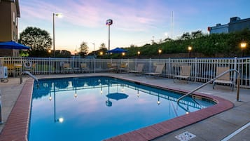A swimming pool with a brick border, surrounded by a white fence, and equipped with lounge chairs and umbrellas.