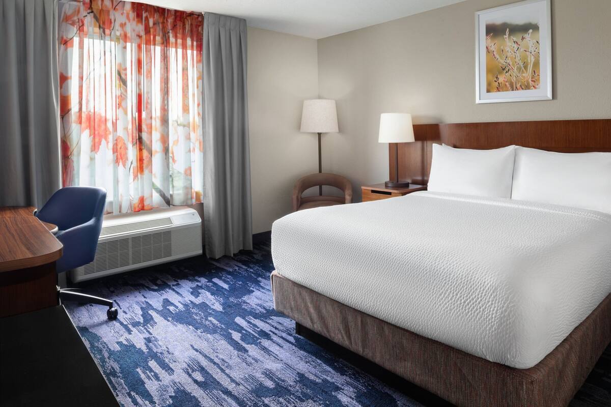 Une chambre d’hôtel avec un grand lit, un bureau avec une chaise, une lampe sur pied et une fenêtre agrémentée de rideaux à motifs floraux.