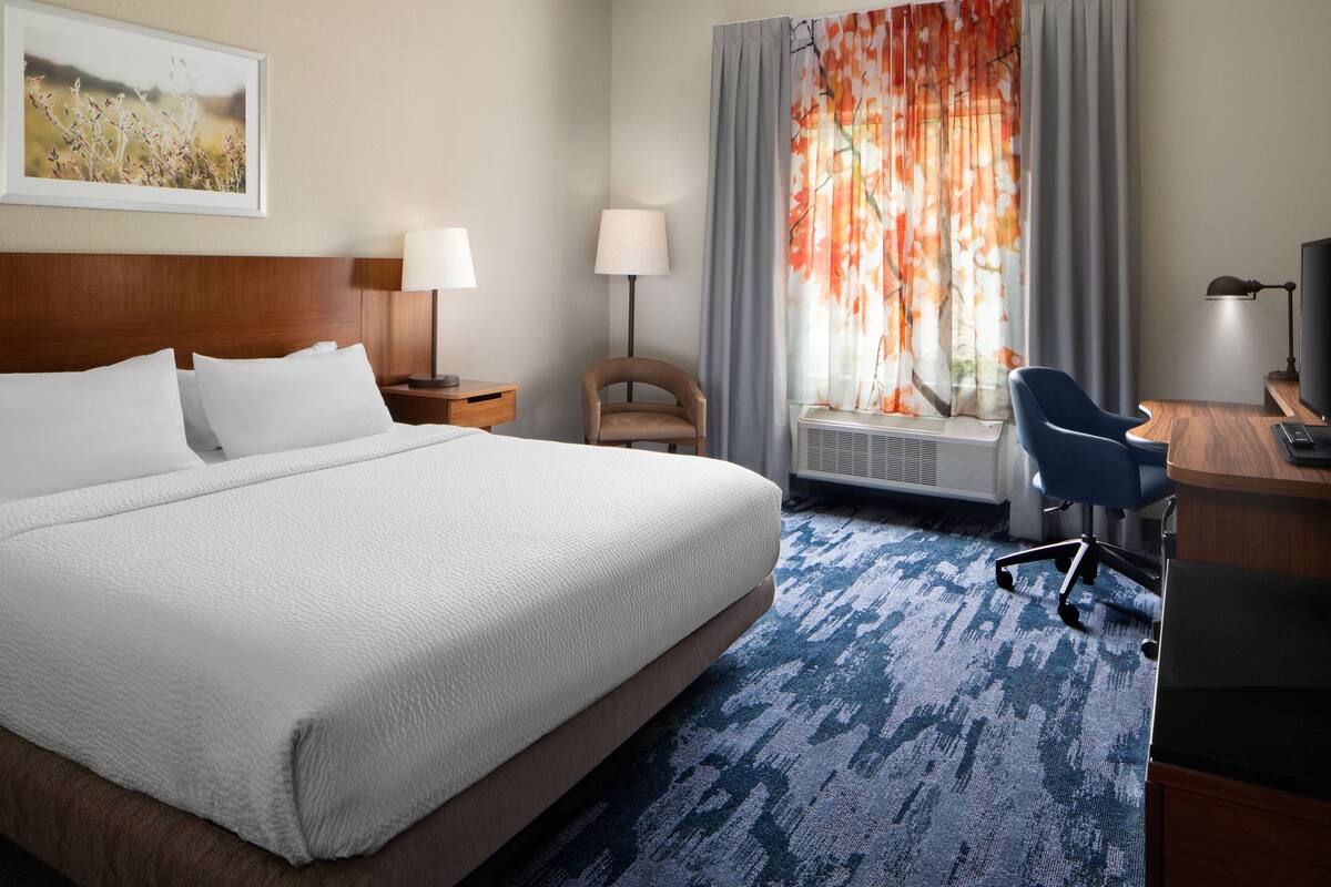 Une chambre d’hôtel avec un grand lit, un bureau, une chaise, une lampe et une fenêtre avec des rideaux.