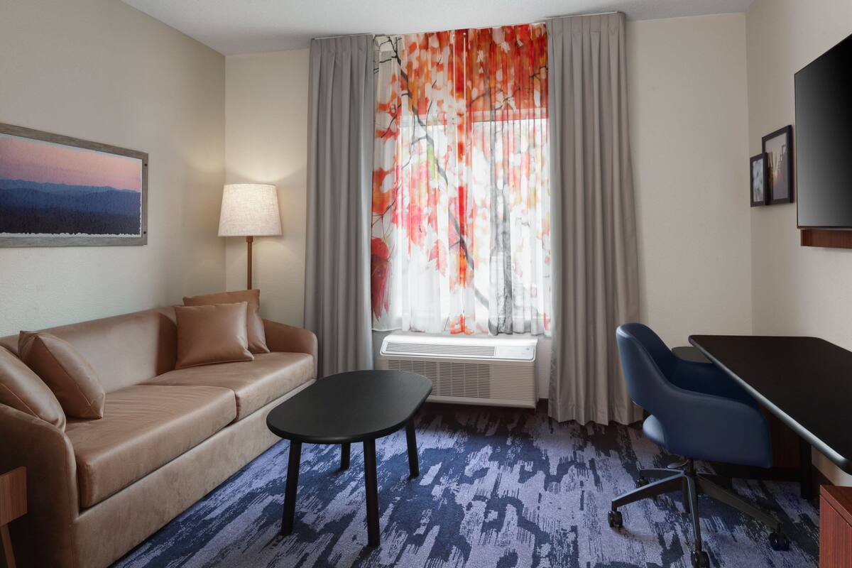 Une chambre d’hôtel équipée d’un canapé, d’une petite table, d’un bureau avec une chaise, d’une fenêtre avec des rideaux et d’un téléviseur fixé au mur.