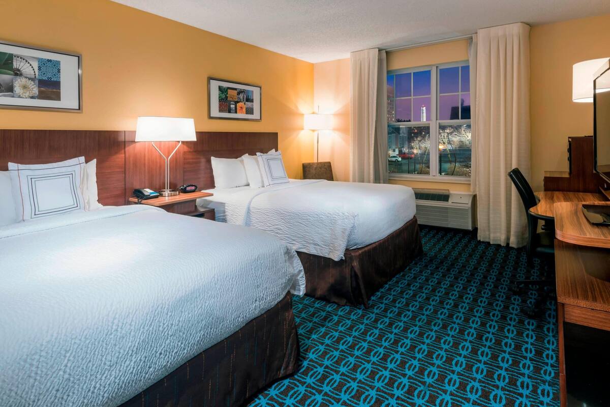 Ein Hotelzimmer mit zwei Betten, einem Schreibtisch, einem Stuhl, einer Lampe, einem Fenster mit Vorhängen und einem gemusterten Teppich.