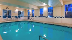 En indendørs swimmingpool med krystalklart blåt vand, et ur på væggen, og et skilt der siger "Ingen livreddere på vagt".
