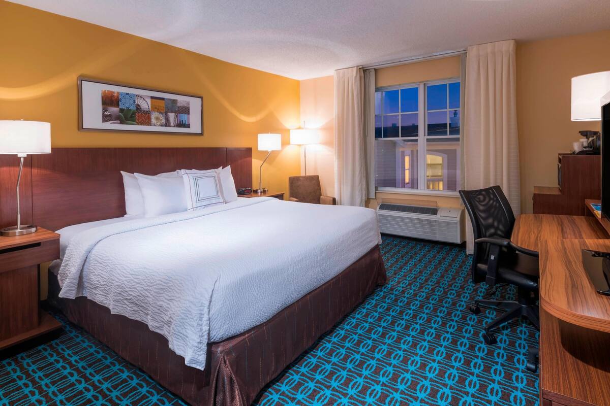 Ein Hotelzimmer mit einem großen Bett, einem Schreibtisch, einem Sessel, einem Fernseher und einem Fenster mit Vorhängen.